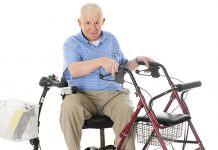 A senior man sitting sideways on his power scooter while holding onto the handles of his wheeling walker. On a white background.