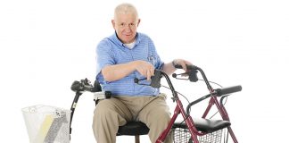 A senior man sitting sideways on his power scooter while holding onto the handles of his wheeling walker. On a white background.