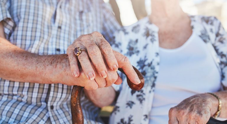 Cropped shot of senior couple holding hands while sitting together. Focus on hands on walking stick.